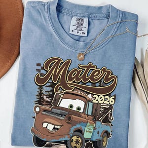 May include: A light blue Comfort Colors t-shirt featuring a graphic of Mater from the movie Cars, with the text "Mater 2026". The shirt has a crew neck and short sleeves. A gold necklace is draped across the shirt.