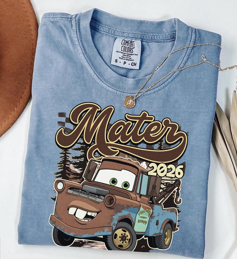 May include: A light blue Comfort Colors t-shirt featuring a graphic of Mater from the movie Cars, with the text "Mater 2026". The shirt has a crew neck and short sleeves. A gold necklace is draped across the shirt.