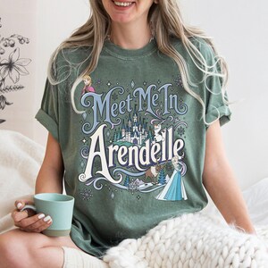 K&ouml;nnte beinhalten: Ein salbeigr&uuml;nes Kurzarm-T-Shirt mit einem Grafikdruck und dem Text "Meet Me In Arendelle". Das Design enth&auml;lt Illustrationen eines Schlosses, Schneeflocken und Figuren wie Anna, Elsa, Olaf und Sven in Blau-, Wei&szlig;- und Braunt&ouml;nen.