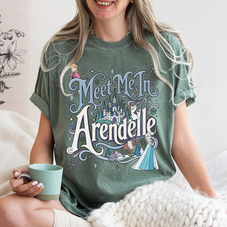 K&ouml;nnte beinhalten: Ein salbeigr&uuml;nes Kurzarm-T-Shirt mit einem Grafikdruck und dem Text "Meet Me In Arendelle". Das Design enth&auml;lt Illustrationen eines Schlosses, Schneeflocken und Figuren wie Anna, Elsa, Olaf und Sven in Blau-, Wei&szlig;- und Braunt&ouml;nen.