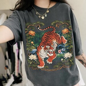 K&ouml;nnte beinhalten: Dunkelgraues T-Shirt mit einer detaillierten Illustration eines Tigers mit orangefarbenen und schwarzen Streifen, umgeben von Blumen und Bl&auml;ttern. Der Tiger steht im Mittelpunkt, vor einem Hintergrund aus Gr&uuml;n- und Blaut&ouml;nen.