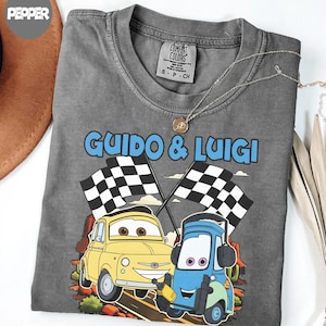 May include: A gray Comfort Colors t-shirt featuring the text "Guido & Luigi" above an illustration of two cartoon cars with checkered flags. The yellow car is next to a blue car. A silver necklace with a pendant is on the shirt.