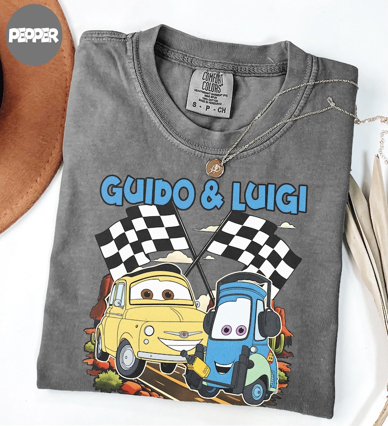 May include: A gray Comfort Colors t-shirt featuring the text "Guido & Luigi" above an illustration of two cartoon cars with checkered flags. The yellow car is next to a blue car. A silver necklace with a pendant is on the shirt.