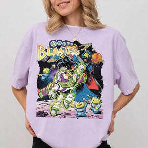 Puede incluir: Una camiseta de color morado claro con un estampado gr&aacute;fico que incluye el texto "ASTRO BLASTERS" en amarillo. El dise&ntilde;o muestra a un ranger espacial con traje blanco y verde, tres peque&ntilde;os alien&iacute;genas verdes y un villano sobre un fondo espacial.