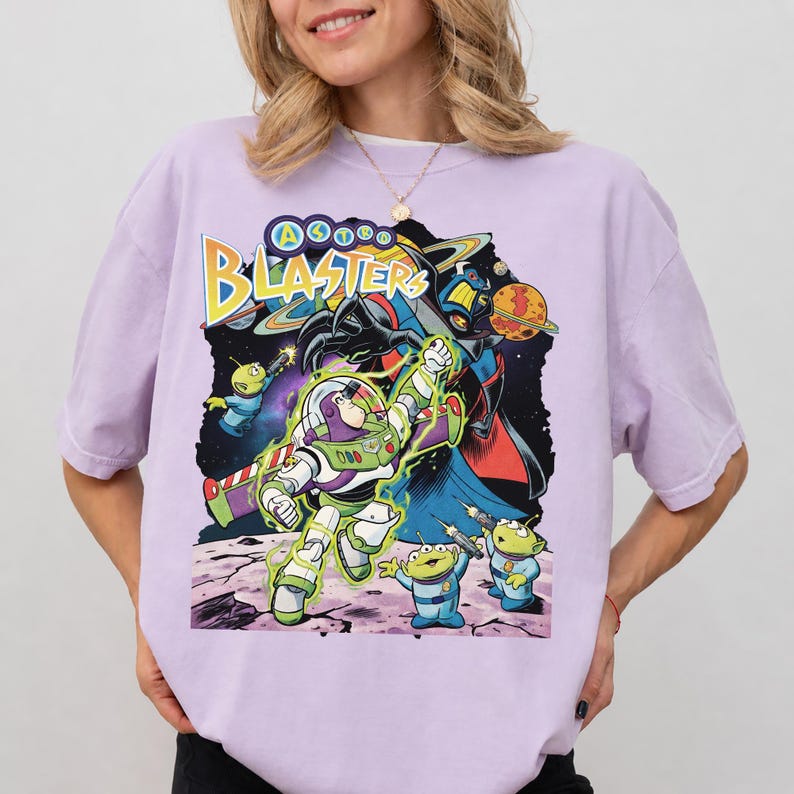 Puede incluir: Una camiseta de color morado claro con un estampado gr&aacute;fico que incluye el texto "ASTRO BLASTERS" en amarillo. El dise&ntilde;o muestra a un ranger espacial con traje blanco y verde, tres peque&ntilde;os alien&iacute;genas verdes y un villano sobre un fondo espacial.