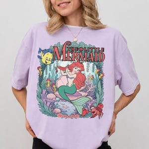 May include: A light purple short-sleeve t-shirt featuring a graphic print of The Little Mermaid with Ariel, Flounder, and Sebastian. The design includes underwater coral, seaweed, and a large seashell, with the text The Little Mermaid above.
