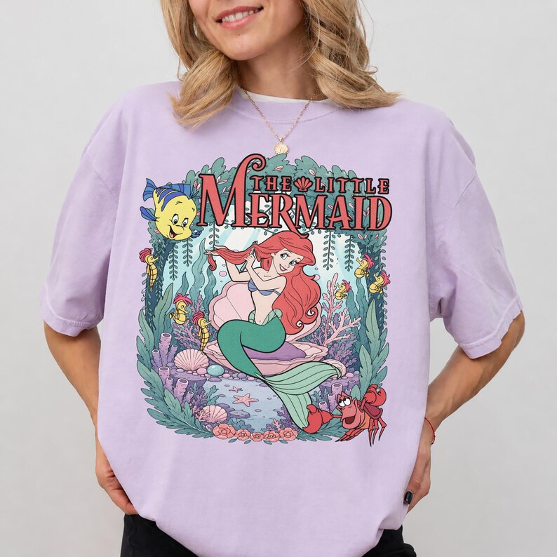 May include: A light purple short-sleeve t-shirt featuring a graphic print of The Little Mermaid with Ariel, Flounder, and Sebastian. The design includes underwater coral, seaweed, and a large seashell, with the text The Little Mermaid above.