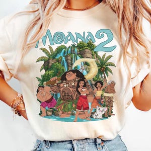 May include: A cream-colored short-sleeve t-shirt featuring a graphic print of the Moana 2 movie characters including Moana, Maui, and Pua. The design includes a tropical waterfall background, palm trees, and the text Moana 2 in light blue letters.