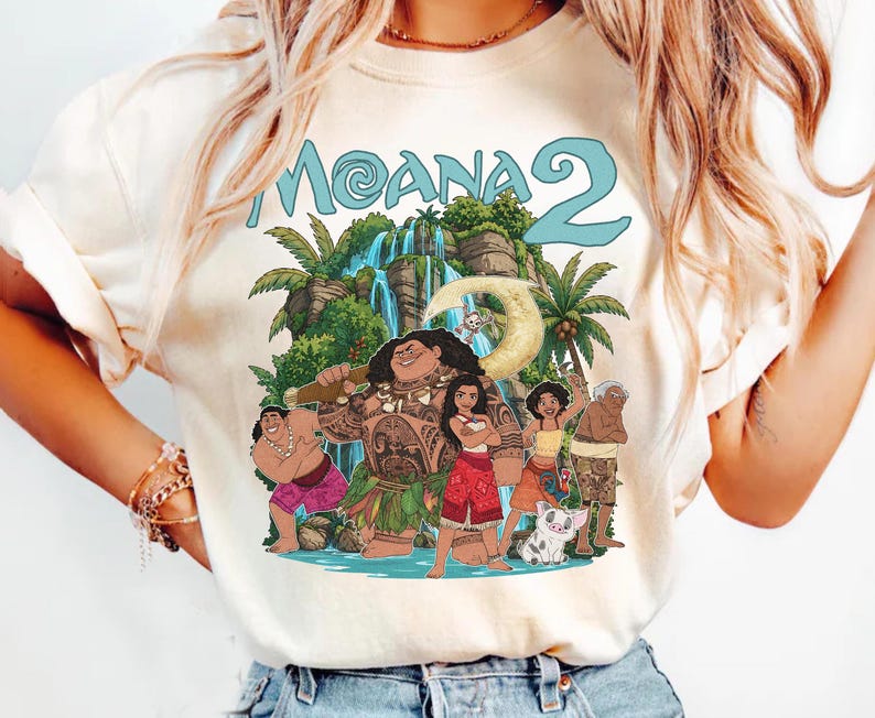 May include: A cream-colored short-sleeve t-shirt featuring a graphic print of the Moana 2 movie characters including Moana, Maui, and Pua. The design includes a tropical waterfall background, palm trees, and the text Moana 2 in light blue letters.
