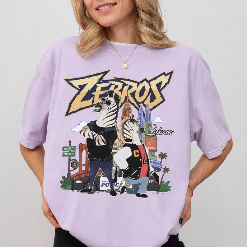 May include: A light purple short-sleeve t-shirt featuring a graphic print of two anthropomorphic zebras in a city setting. The text "ZEBROS" appears in large yellow letters at the top, with "Zootopia" and "Hopps" street signs visible in the background.