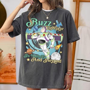 May include: A charcoal gray t-shirt featuring a graphic of Buzz Lightyear with the text Buzz Lightyear To Infinity And Beyond. The design includes a blue swirl background, planets, a satellite, and a comet with yellow stars on the shirt front.