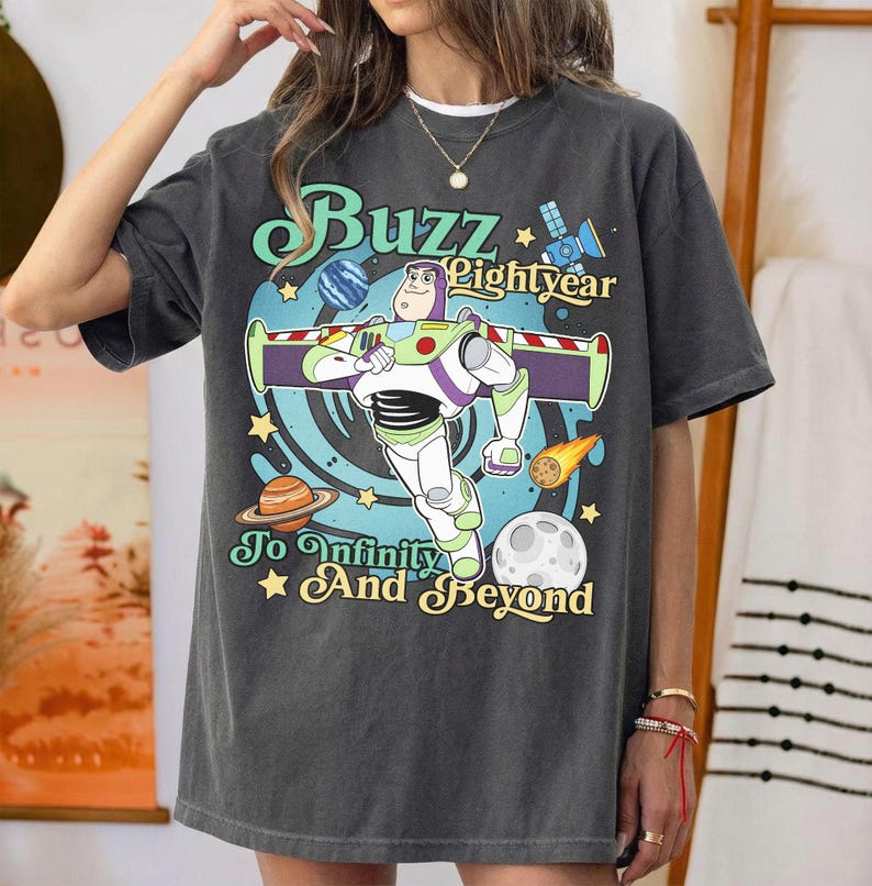 May include: A charcoal gray t-shirt featuring a graphic of Buzz Lightyear with the text Buzz Lightyear To Infinity And Beyond. The design includes a blue swirl background, planets, a satellite, and a comet with yellow stars on the shirt front.