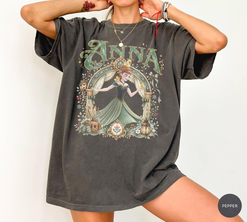 May include: A charcoal gray short-sleeve t-shirt featuring a graphic print of the character Anna from Frozen. The design includes the name Anna in green lettering, a decorative archway, floral elements, a small bird, and a lantern.