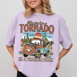 May include: A lilac t-shirt with a cartoon tow truck design and the text "Happier than a Tornado in a Trailer Park." The graphic includes a desert scene with cacti and a setting sun. The shirt has a loose fit.