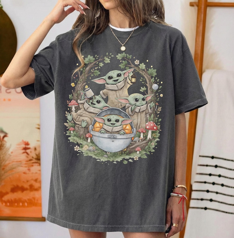 May include: A charcoal gray short-sleeve t-shirt featuring a graphic print of four small green creatures with large ears. The illustration shows them in a forest setting with mushrooms, foliage, and one creature sitting inside a metallic pod.