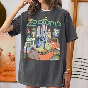 May include: A charcoal gray short-sleeve t-shirt featuring a graphic print of the characters Judy Hopps and Nick Wilde from Zootopia. The design includes the text Zootopia and Bunny Best Friend, set against a colorful city skyline background.