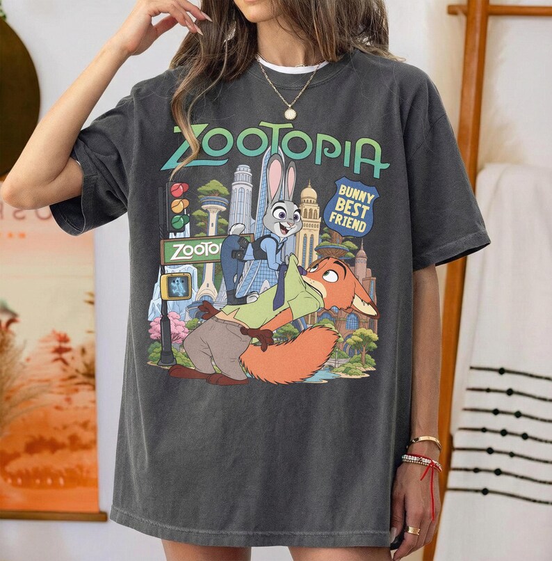 May include: A charcoal gray short-sleeve t-shirt featuring a graphic print of the characters Judy Hopps and Nick Wilde from Zootopia. The design includes the text Zootopia and Bunny Best Friend, set against a colorful city skyline background.