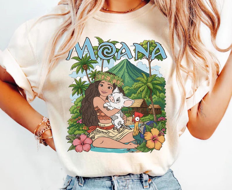 May include: A cream-coloured t-shirt featuring a graphic print of the character Moana holding a piglet, with a rooster, tropical palm trees, a mountain, and the text MOANA in light blue letters at the top, surrounded by colourful hibiscus flowers.