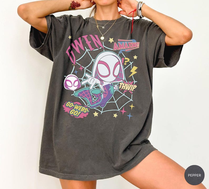 May include: A charcoal gray short-sleeve t-shirt featuring a graphic of a cartoon character in a white and pink mask with a spiderweb background. The print includes the text GWEN, AMAZING, THWIP, and GO-WEBS-GO in colorful, stylized lettering.