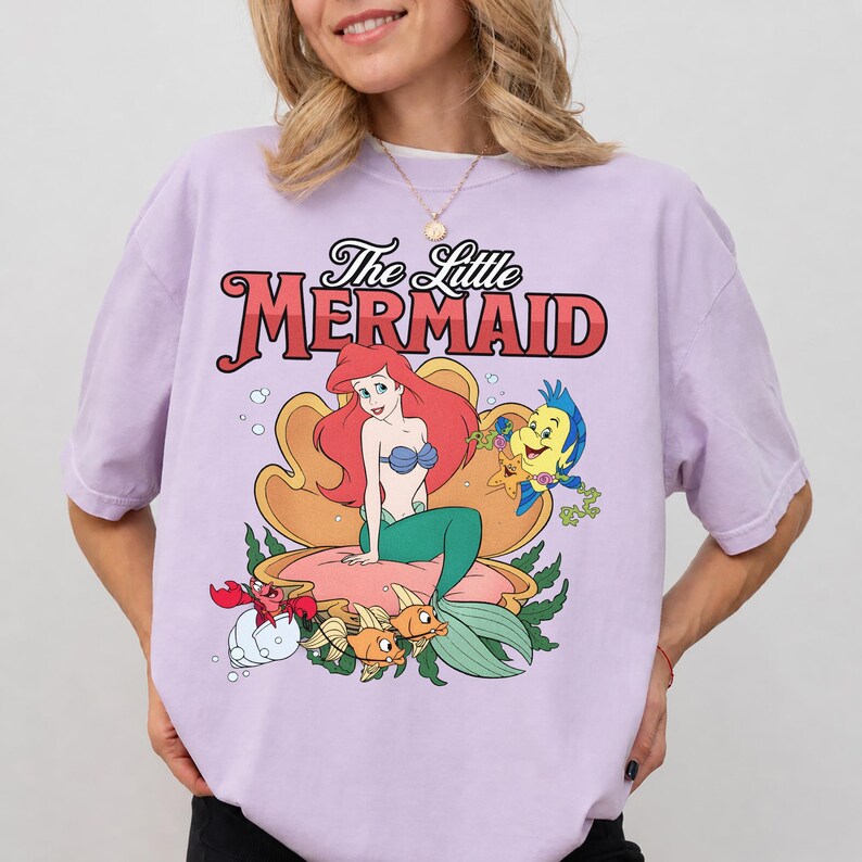 May include: A light purple t-shirt featuring a graphic print of The Little Mermaid with Ariel sitting in a large seashell, accompanied by Flounder, Sebastian, and other sea creatures. The text The Little Mermaid is printed in red and black.