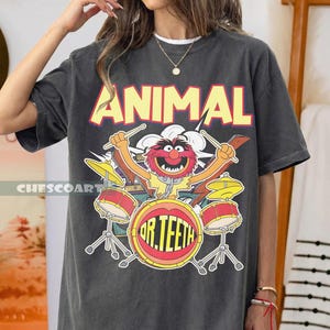 Könnte beinhalten: Dunkelgraues T-Shirt mit einem Cartoon-Bild von Animal von den Muppets, der Schlagzeug spielt. Das Wort "ANIMAL" steht in gelben Blockbuchstaben über der Figur. Auf dem Schlagzeug steht "DR. TEETH" auf der Bassdrum.