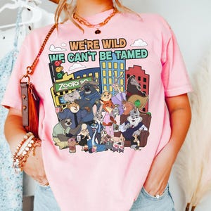 May include: Pink t-shirt featuring a colorful graphic of characters from the movie Zootopia, with the text "WE'RE WILD WE CAN'T BE TAMED". The design includes a cityscape and traffic lights.