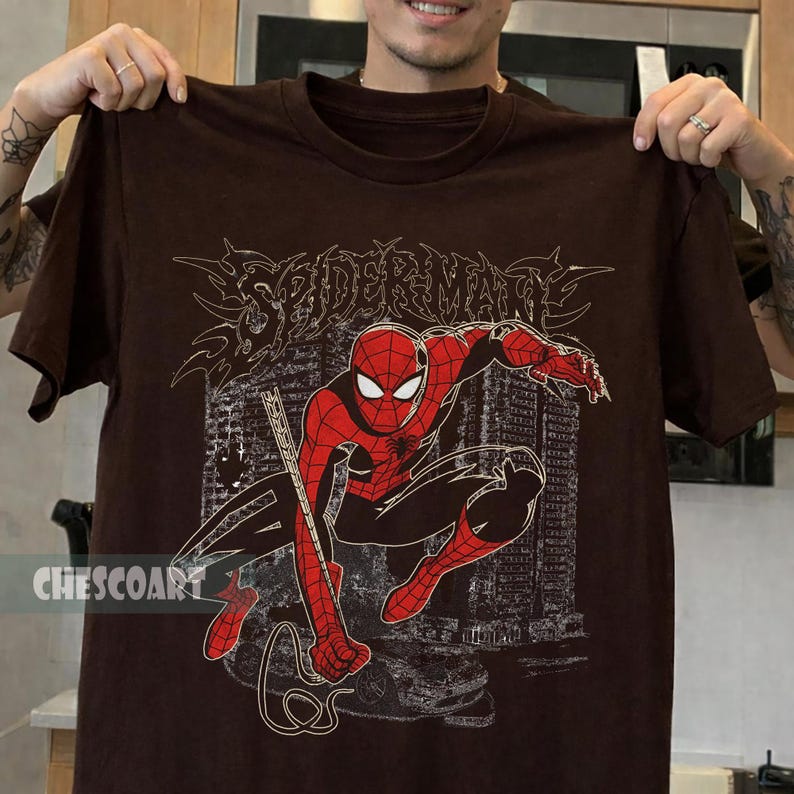 May include: A brown t-shirt featuring a graphic of Spider-Man in a dynamic pose, with a cityscape backdrop. The design includes the word "SPIDERMAN" in a stylized font. The brand name "CHESCOART" is printed on the bottom left.