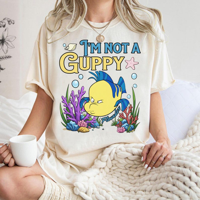 Puede incluir: Camiseta color crema con un personaje de dibujos animados de Flounder y el texto "I'M NOT A GUPPY". El dise&ntilde;o incluye colores azul, amarillo y morado, con burbujas y detalles de coral. La camiseta es de corte holgado.