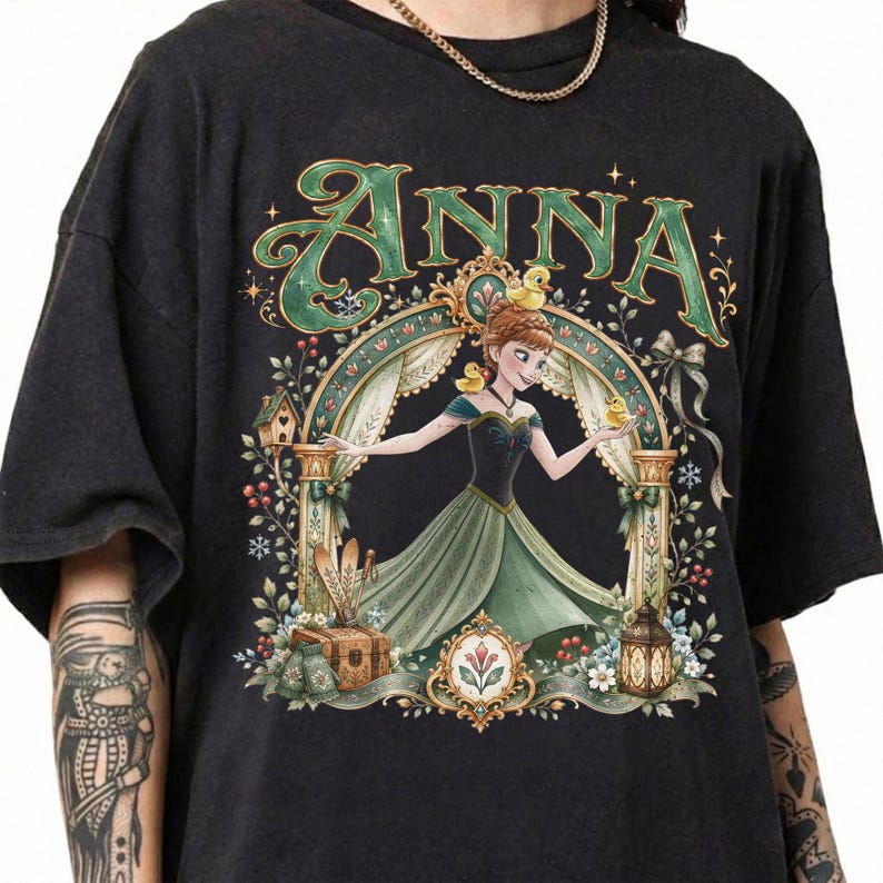 May include: A black short-sleeve t-shirt featuring a graphic print of a character named Anna in a green dress, surrounded by an ornate archway, small yellow ducks, a wooden chest, a lantern, and floral elements with the text ANNA at the top.