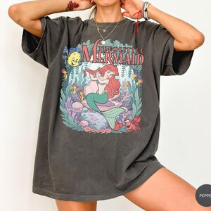 May include: A charcoal gray short-sleeve t-shirt featuring a graphic print of The Little Mermaid with Ariel, Flounder, and Sebastian. The design includes underwater elements like coral, seaweed, and seashells, with the text The Little Mermaid.
