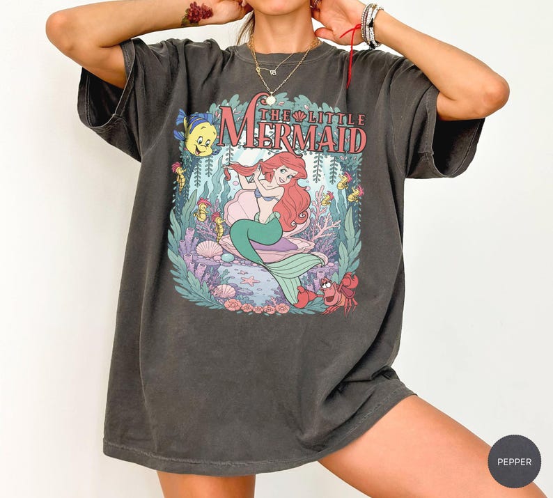 May include: A charcoal gray short-sleeve t-shirt featuring a graphic print of The Little Mermaid with Ariel, Flounder, and Sebastian. The design includes underwater elements like coral, seaweed, and seashells, with the text The Little Mermaid.