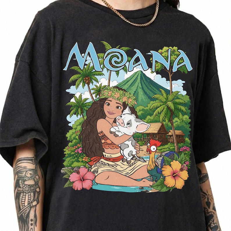 May include: A black t-shirt featuring a graphic print of the character Moana holding a piglet, with a rooster standing nearby. The background shows a tropical island landscape with palm trees, a mountain, and huts. The word MOANA is printed in blue.