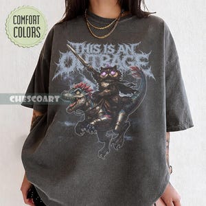 May include: Dark gray t-shirt featuring a graphic of a cat riding a dinosaur and holding a sword, with the text "THIS IS AN OUTRAGE" in a stylized font. The shirt also has the text "CHESCOART" and "COMFORT COLORS".