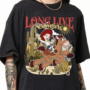 Puede incluir: Una camiseta negra con un estampado gr&aacute;fico de una vaquera de dibujos animados montando un caballo marr&oacute;n con un lazo. El texto "LONG LIVE cowgirls" aparece sobre la ilustraci&oacute;n, que incluye cactus, un sol y plantas del desierto en tonos apagados.