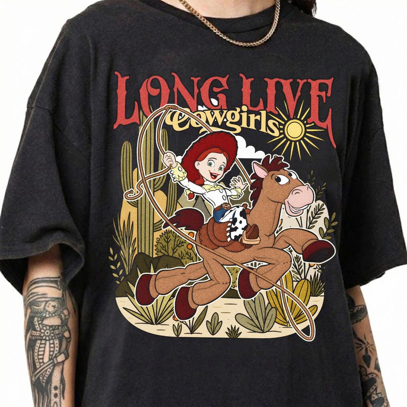Puede incluir: Una camiseta negra con un estampado gr&aacute;fico de una vaquera de dibujos animados montando un caballo marr&oacute;n con un lazo. El texto "LONG LIVE cowgirls" aparece sobre la ilustraci&oacute;n, que incluye cactus, un sol y plantas del desierto en tonos apagados.
