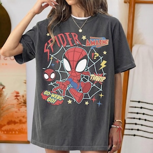 May include: A charcoal gray short-sleeve t-shirt featuring a graphic of a cartoon Spider-Man character centered over a white spiderweb design. The print includes the red text "SPIDER", "AMAZING!", "THWIP", and "GO-WEBS-GO!" along with yellow lightning bolts and stars.