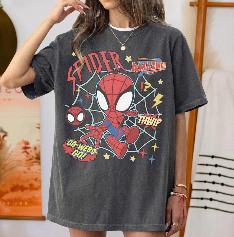 May include: A charcoal gray short-sleeve t-shirt featuring a graphic of a cartoon Spider-Man character centered over a white spiderweb design. The print includes the red text "SPIDER", "AMAZING!", "THWIP", and "GO-WEBS-GO!" along with yellow lightning bolts and stars.