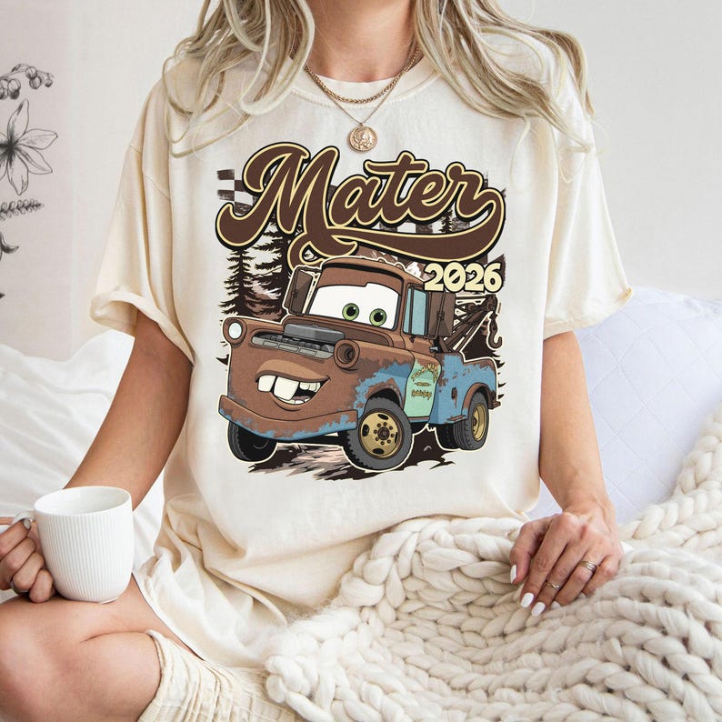 May include: Cream-colored t-shirt featuring a cartoon image of Mater from the movie Cars. The graphic includes the word "Mater" in brown script, the year "2026", and a forest backdrop. The tow truck character is brown and blue.