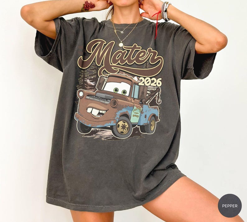 May include: Dark gray t-shirt featuring a graphic of Mater from the movie Cars, with the text "Mater 2026" in brown and white. The design includes a cartoon image of the tow truck Mater. The shirt has short sleeves and a relaxed fit.