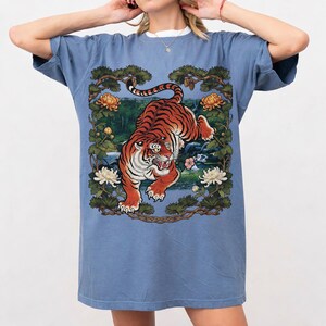 K&ouml;nnte beinhalten: Ein blaues T-Shirt mit einer detaillierten Illustration eines Tigers mit orangefarbenen und schwarzen Streifen, vor einem Hintergrund aus Blumen und Bl&auml;ttern. Der Tiger ist der Mittelpunkt, in einer dynamischen Pose. Das Shirt hat kurze &Auml;rmel und eine lockere Passform.