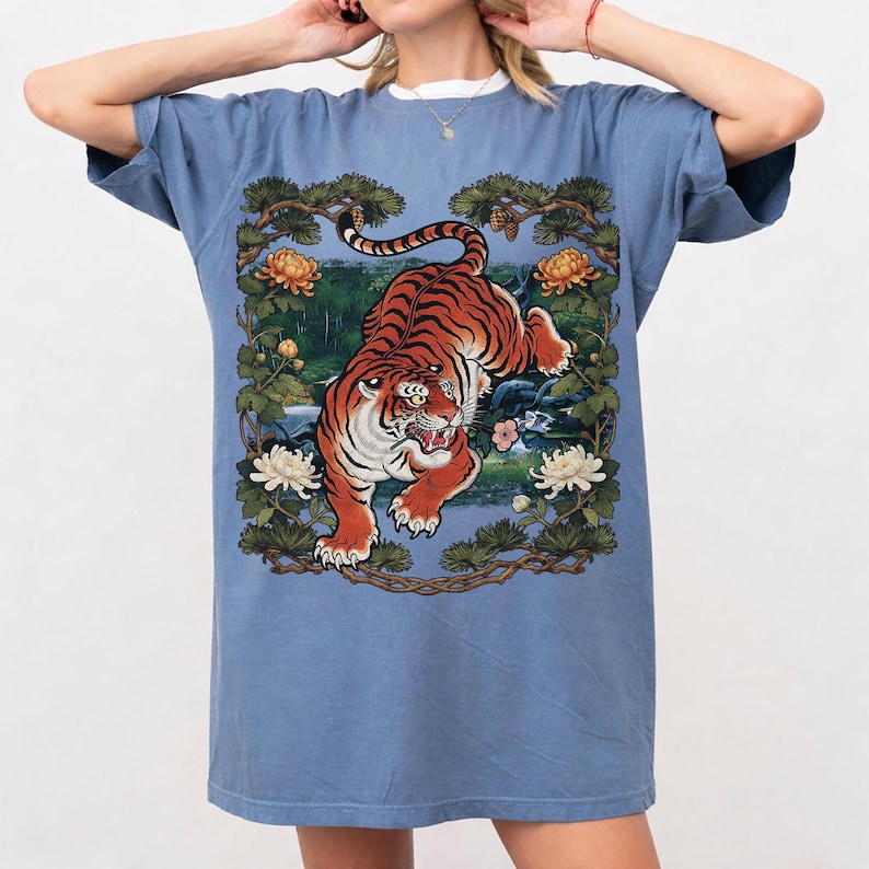 K&ouml;nnte beinhalten: Ein blaues T-Shirt mit einer detaillierten Illustration eines Tigers mit orangefarbenen und schwarzen Streifen, vor einem Hintergrund aus Blumen und Bl&auml;ttern. Der Tiger ist der Mittelpunkt, in einer dynamischen Pose. Das Shirt hat kurze &Auml;rmel und eine lockere Passform.