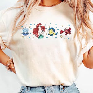 K&ouml;nnte beinhalten: Cremefarbenes T-Shirt mit einem Aquarelldesign von Figuren aus einem beliebten Zeichentrickfilm. Das Design umfasst eine Perle, eine Meerjungfrau mit roten Haaren, einen gelb-blauen Fisch und eine rote Krabbe mit blauen Punkten und gelben Sternen.