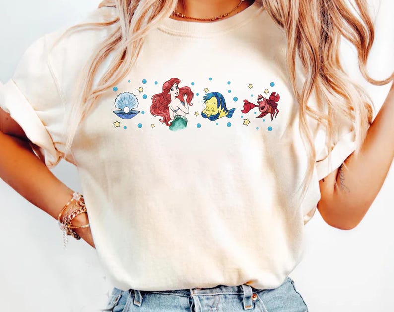 K&ouml;nnte beinhalten: Cremefarbenes T-Shirt mit einem Aquarelldesign von Figuren aus einem beliebten Zeichentrickfilm. Das Design umfasst eine Perle, eine Meerjungfrau mit roten Haaren, einen gelb-blauen Fisch und eine rote Krabbe mit blauen Punkten und gelben Sternen.