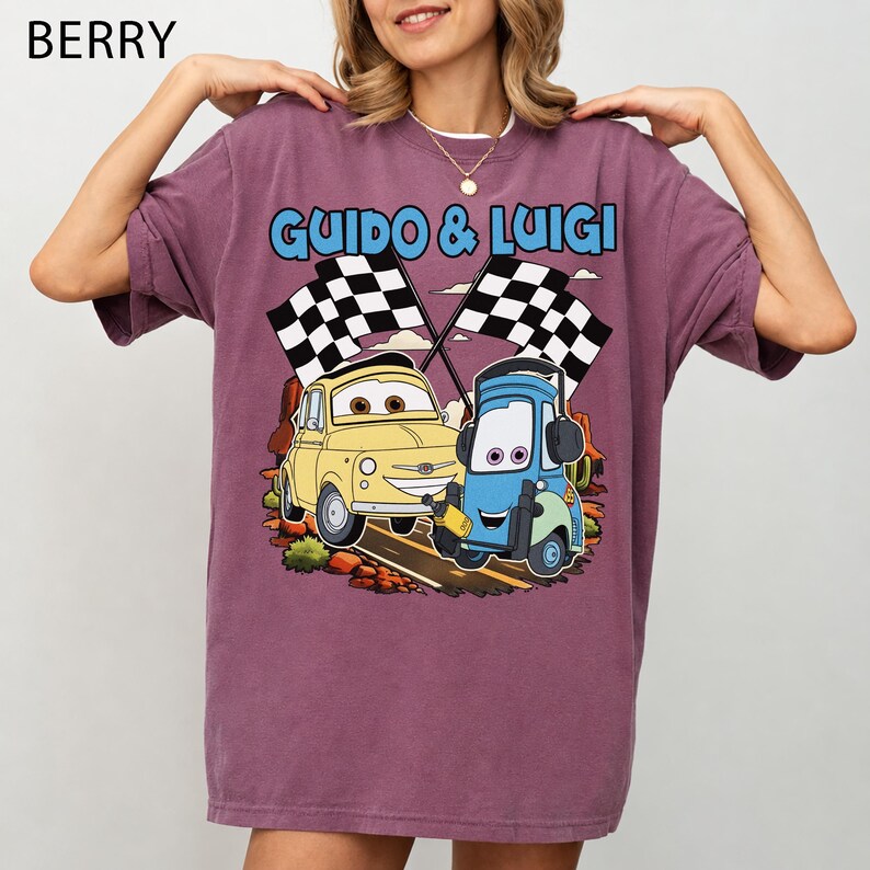 May include: Berry-colored t-shirt featuring a cartoon graphic of two cars, Guido and Luigi, with checkered flags. The text "GUIDO & LUIGI" is above the cars. The shirt has short sleeves and a relaxed fit.