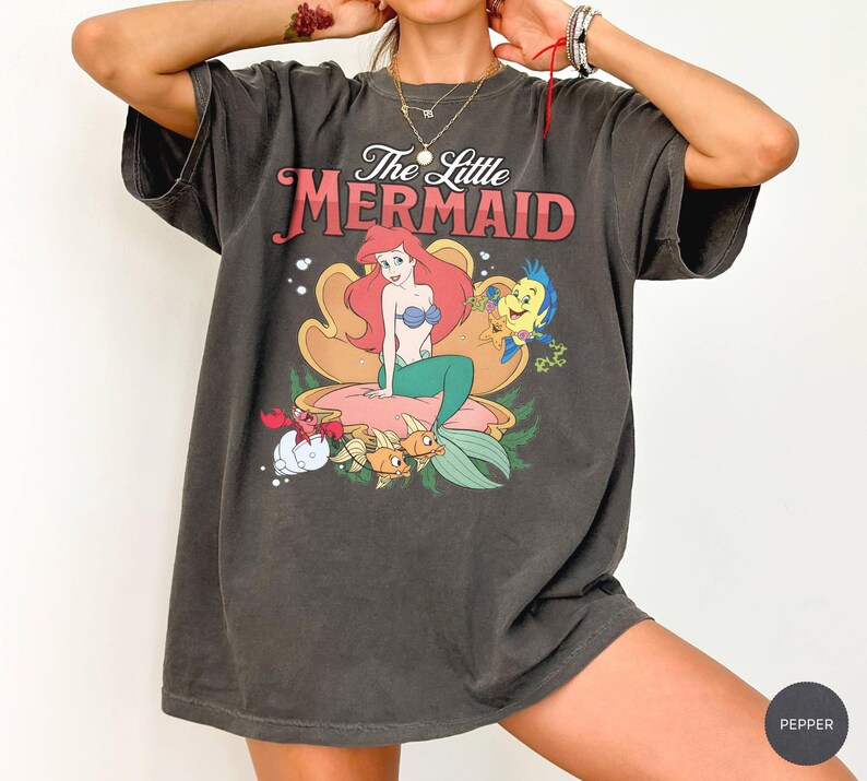 May include: A charcoal gray short-sleeve t-shirt featuring a graphic print of The Little Mermaid with Ariel, Flounder, and Sebastian. The design shows Ariel sitting in a large seashell with the text The Little Mermaid printed in red and white.