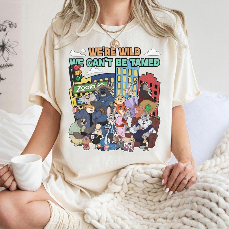 May include: Cream-colored t-shirt featuring a colorful graphic of characters from the animated movie "Zootopia" with the text "WE'RE WILD WE CAN'T BE TAMED". The design includes a cityscape and various animal characters.
