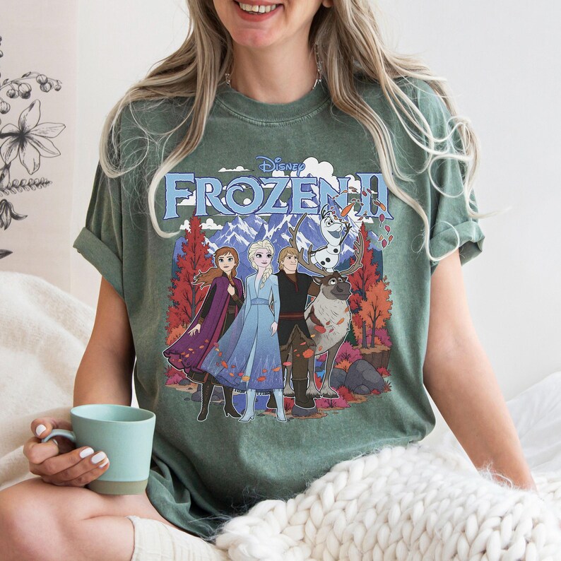 May include: A sage green short-sleeve t-shirt featuring a graphic print of the Disney Frozen II logo with characters Anna, Elsa, Kristoff, Olaf, and Sven standing in a forest setting with mountains in the background and falling autumn leaves.
