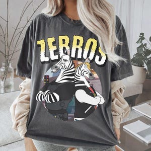 Puede incluir: Camiseta gris oscuro con la palabra "ZEBROS" en letras de bloque amarillas y blancas. La camiseta presenta una ilustración de dibujos animados de dos cebras con los brazos cruzados, que llevan chaquetas. Las cebras están sobre un fondo circular.