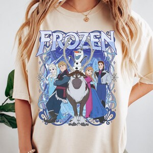 May include: A cream-colored short-sleeve t-shirt featuring a large graphic print of the Disney Frozen characters Elsa, Kristoff, Olaf, Sven, Anna, and Hans. The design includes the word FROZEN in large blue letters with intricate blue line art.