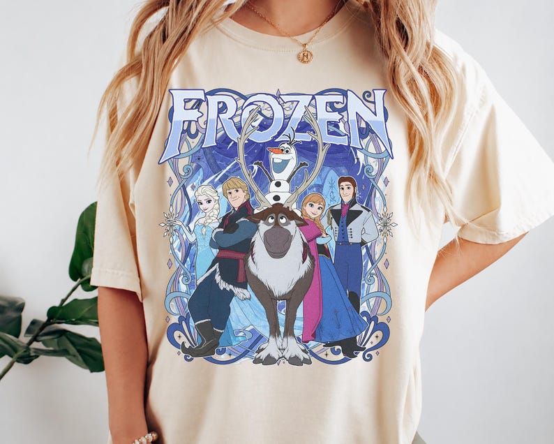 May include: A cream-colored short-sleeve t-shirt featuring a large graphic print of the Disney Frozen characters Elsa, Kristoff, Olaf, Sven, Anna, and Hans. The design includes the word FROZEN in large blue letters with intricate blue line art.